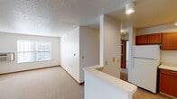 This single-floor unit offers a functional, well-organized layout designed for comfortable everyday living. The open living area is finished with neutral-toned carpet, white walls, and a textured ceiling, creating a clean and understated aesthetic throughout. Natural light enters through blinded windows, and baseboard heating along with a wall-mounted HVAC unit ensures year-round climate control.

The kitchen is a standout feature, equipped with rich medium-toned wood cabinetry extending to the upper walls for ample storage. White appliances—including a refrigerator, electric range, and dishwasher—complement the light countertops. A pass-through counter connects the kitchen to the living area, enhancing the sense of openness. Recessed and flush-mount lighting illuminates the space effectively.

The bedroom is generously sized, featuring carpet flooring, a textured ceiling, and direct access to a walk-in closet for added storage convenience. A hallway connects the bedroom to the rest of the unit.

The bathroom is thoughtfully designed with accessibility in mind, featuring grab bars in the shower/tub area, a single-sink vanity with warm wood cabinetry, a mirror, and tile flooring. The layout is spacious and easy to navigate throughout.
