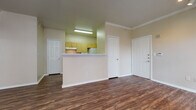 This single-story unit is finished consistently throughout with dark, wood-style vinyl plank flooring and neutral beige walls, creating a cohesive and practical aesthetic across all spaces. Crown molding accents the living area and select rooms, adding a subtle architectural detail to the otherwise straightforward design.

The open living area is well-proportioned, featuring track lighting and direct sightlines to the kitchen, which is separated by a half-wall peninsula. The kitchen is laid out in a galley configuration with light wood cabinetry, white laminate countertops, and white appliances including a refrigerator, dishwasher, and microwave. An electrical panel is accessible via a dedicated utility closet off the kitchen.

The unit includes multiple bedrooms, each with wood-style flooring, window blinds for natural light control, and individual closets. One bedroom features a walk-in closet, while another includes a ceiling fan and crown molding. Two bathrooms serve the unit: one with a tub/shower combination, single vanity, and light wood cabinetry, and a second more compact bathroom with a shower and basic fixtures. Both bathrooms continue the wood-style flooring found throughout the unit.