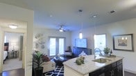 This single-floor residence showcases an open-concept layout that connects the kitchen and living areas into one cohesive, functional space. The kitchen features striking granite countertops with a generous island that serves as a natural dividing point between the cooking and living zones. A stainless steel sink is integrated into the island, and pendant lights hang above, adding focused illumination and a modern aesthetic touch.

The living area is anchored by a plush sofa and accented with a bold navy and white geometric-patterned area rug, creating a stylish focal point. A ceiling fan with an integrated light fixture serves both the practical and visual needs of the space. Wall-mounted decorative metallic pieces add an artistic element to the neutral walls.

Natural light filters in through multiple windows fitted with blinds, brightening the interior throughout the day. French doors along one wall offer additional light and potential outdoor access. A media console with a flat-screen television grounds the entertainment area. Lush indoor plants add warmth and life to the space, contributing to an inviting and well-balanced atmosphere throughout.