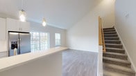 Located within the Cornerstone at Hauppauge community at 135 Stonehenge Lane, this two-floor residence offers a well-designed layout with a clean, contemporary aesthetic throughout. The main living area features vaulted ceilings that lend an airy, open feel, complemented by large windows that invite an abundance of natural light. Grey-toned hardwood-style flooring runs through the main living and kitchen areas, adding warmth and cohesion to the space. The kitchen is appointed with crisp white shaker-style cabinetry, stainless steel appliances including a gas range, microwave, and French door refrigerator, along with light quartz countertops and a subway tile backsplash. A sliding glass door off the kitchen opens to an outdoor balcony, seamlessly connecting indoor and outdoor living. Carpeted bedrooms provide comfort and quiet, each with generous windows overlooking the surrounding community. The upper level features an open loft-style area with recessed lighting and plush carpeting, offering flexible use as a lounge or workspace. A carpeted staircase with a wood handrail connects both floors. The bathrooms feature tile flooring and dark vanity cabinetry with light countertops, delivering a stylish contrast. Throughout, the residence maintains a bright, neutral palette that creates a welcoming and versatile living environment.