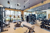 Located at 1505 Cornerstone Blvd in Daytona Beach, FL, this fitness center offers a thoughtfully designed exercise space that balances functionality with modern aesthetics. The single-floor layout provides a generous workout area filled with a comprehensive selection of Matrix-brand cardio equipment, including treadmills, ellipticals, and stationary bikes. The strength training zone is equally well-appointed, featuring a multi-functional cable machine, adjustable benches, a full dumbbell rack, kettlebells, and stability balls.

The room's design is a standout feature, with warm wood-paneled walls and a bold dark ceiling that create a sleek, contemporary atmosphere. Stylish cylindrical pendant lights hang throughout, adding focused illumination. A large floor-to-ceiling window spans one wall, flooding the space with natural light and offering outdoor views of the surrounding community and landscaping. Full-length mirrors on the opposite wall visually expand the room while serving a practical purpose for form-checking during workouts.

Coastal-themed wall murals add a distinctive local touch, tying the space to its Florida setting. The neutral carpeted flooring provides comfort underfoot, and the overall ambiance is both motivating and inviting — making this fitness center a standout amenity.
