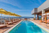 Located in the heart of San Jose, CA, this rooftop pool deck offers an elevated outdoor retreat with sweeping panoramic views of the city skyline and the surrounding mountain ranges. The centerpiece of the space is a generously sized lap pool with crystal-clear water, framed by large-format terracotta-toned tiles laid in a classic checkered pattern that adds a refined, resort-like aesthetic throughout the deck.

Along the poolside, rows of striped sun loungers are shaded by a series of bold yellow scalloped umbrellas, creating a vibrant and inviting atmosphere ideal for relaxation. A separate hot tub area provides an additional aquatic amenity, positioned to take full advantage of the unobstructed views.

The lounge zone features comfortable outdoor seating arrangements, including a curved white sofa set beneath a covered terrace supported by exposed concrete columns — offering a shaded retreat with glass railings that preserve the scenic vistas. Warm wood-paneled walls on the service building add a natural, contemporary contrast to the concrete and tile surroundings, while large glass sliding doors provide access to interior facilities.

Potted tropical plants and greenery are thoughtfully placed throughout the deck, softening the urban setting and enhancing the overall ambiance. The open-sky exposure and elevated position ensure abundant natural light, making this rooftop an exceptional amenity space.