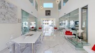 This co-share work table is situated within a beautifully designed communal space that combines modern aesthetics with a bright, open atmosphere. The area is defined by its soaring double-height ceilings that amplify the sense of scale and openness, while polished white marble flooring with subtle grey veining reflects abundant natural light throughout the space.

Floor-to-ceiling glass partitions line both sides of the central corridor, offering transparent views into adjacent work and lounge areas while maintaining a sense of connectivity. Large sliding glass doors at the far end open directly to an outdoor pool area, flooding the interior with natural light and providing an inviting visual connection to the exterior.

The co-share work table itself is a sleek, minimalist white surface accompanied by transparent acrylic ghost chairs, contributing to the space's clean, contemporary feel. Decorative sputnik chandeliers hang overhead, complementing the sculptural wall art and organic branch installations that add artistic character. Modern white lounge seating with bold red accent pillows is strategically placed throughout, creating comfortable breakout areas. The overall ambiance is crisp, professional, and energizing—ideal for focused, collaborative work.