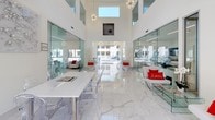 This co-share work table is situated within a beautifully designed communal space that combines modern aesthetics with a bright, open atmosphere. The area is defined by its soaring double-height ceilings that amplify the sense of scale and openness, while polished white marble flooring with subtle grey veining reflects abundant natural light throughout the space.

Floor-to-ceiling glass partitions line both sides of the central corridor, offering transparent views into adjacent work and lounge areas while maintaining a sense of connectivity. Large sliding glass doors at the far end open directly to an outdoor pool area, flooding the interior with natural light and providing an inviting visual connection to the exterior.

The co-share work table itself is a sleek, minimalist white surface accompanied by transparent acrylic ghost chairs, contributing to the space's clean, contemporary feel. Decorative sputnik chandeliers hang overhead, complementing the sculptural wall art and organic branch installations that add artistic character. Modern white lounge seating with bold red accent pillows is strategically placed throughout, creating comfortable breakout areas. The overall ambiance is crisp, professional, and energizing—ideal for focused, collaborative work.