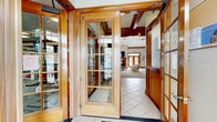 Located at 231 Windmill Ct in Phillipsburg, NJ, this well-appointed clubhouse spans a generous single floor designed to serve as a full-featured community gathering space. The entrance greets visitors with rich warm woodwork, elegant glass-paneled French doors, leaded glass sidelights, and large-format tile flooring, setting a refined tone throughout. The expansive main lounge is the heart of the facility, featuring soaring vaulted ceilings with exposed wood beams, ceiling fans, decorative pendant lighting, a wood-trimmed fireplace, wall-mounted television, and plush carpeting — all creating a welcoming, comfortable atmosphere. Adjacent areas include a game room with abundant natural light from multiple windows and space for recreational activities such as foosball. A fully equipped kitchen provides stainless steel appliances, rich wood cabinetry, granite countertops, and tile flooring suited for event hosting. The dedicated fitness center, accessed through glass-paned French doors, offers a wide array of strength and cardio equipment, mirrored walls, and durable patterned carpet flooring. A private office or conference room with a round table provides a quiet meeting space. Two full bathrooms feature tile flooring, granite vanities, and shower facilities. The hallway connects all spaces seamlessly, completing this comprehensive and functional clubhouse.