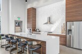 Located at 10568 Gateway Promenade in El Monte, CA, this single-floor community space offers a generously sized, open-concept layout designed with modern aesthetics and functional versatility in mind. The soaring double-height ceilings create an impressive sense of volume, while floor-to-ceiling glass walls and large-pane windows flood the interior with natural light and provide expansive views of the surrounding outdoor courtyard and patio areas.

The kitchen is outfitted with rich wood-grain cabinetry, a textured sculptural backsplash, stainless steel appliances including a refrigerator and range with hood, and a long, white quartz island with bar seating — ideal for both casual use and social gatherings. Overhead, a striking slatted wood ceiling feature and an artistic pendant light add warmth and visual interest to the space.

The living area is thoughtfully appointed with contemporary lounge furniture, a billiards table, and flexible seating arrangements. A decorative wood slat partition serves as an architectural focal point, complementing the polished concrete floors throughout. The hallway connects to additional areas and is accented with framed artwork along the walls. Large sliding glass doors seamlessly extend the interior to an outdoor terrace, enhancing the overall sense of openness and connectivity.