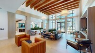 Located at 375 Summer Cypress Cove in Longwood, Florida, this single-floor community amenity space offers a sophisticated and inviting atmosphere defined by thoughtful design and high-end finishes throughout. The expansive main lounge serves as the centerpiece of the space, featuring soaring ceilings with striking exposed wood beam detailing, light hardwood flooring, and floor-to-ceiling black-framed windows that flood the interior with abundant natural light and provide views of the outdoor surroundings. Plush seating arrangements, including channeled leather armchairs in warm cognac tones and a teal sofa accented with coastal-inspired throw pillows, create comfortable social zones throughout. Pendant lighting with woven shades adds warmth and texture to the airy volume above. Adjacent to the lounge is a well-appointed catering kitchen featuring quartz countertops, stainless steel appliances, a decorative textured ceiling treatment, and teal tile backsplash accents that bring a lively pop of color to the space. A separate nook area offers intimate booth-style seating with bold yellow upholstery and circular mirrors, ideal for smaller gatherings or casual conversations. Built-in wood shelving and millwork details throughout add warmth and architectural interest, completing the cohesive, modern coastal aesthetic that defines this community gathering space.