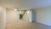 This three-floor home offers a practical and well-organized layout designed to accommodate a variety of living needs. The main living area is finished with wall-to-wall carpeting that adds warmth and comfort underfoot, while neutral-toned walls provide a clean, versatile backdrop throughout. A ceiling fan with an integrated light fixture is centrally positioned, contributing to both airflow and ambient lighting in the space.

A central staircase with a sleek metal railing serves as the visual focal point of the main level, connecting all three floors and creating a sense of vertical continuity. The stairwell is flanked by doorways that lead to additional rooms, suggesting a thoughtfully compartmentalized floor plan. Transom-style windows above the doorways allow natural light to filter through, brightening the interior even in areas away from exterior walls.

Crown molding adds a subtle architectural detail along the ceiling line, lending a polished finish to the space. The overall design maintains a straightforward, functional aesthetic with clean lines and a neutral palette, making it highly adaptable to a range of furnishing styles. With multiple levels to explore, this home provides ample space for both everyday living and flexible use across its various rooms and areas.