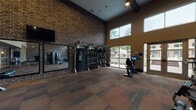 This fitness center is designed to accommodate a wide range of workout styles within a spacious, single-floor layout. The architecture is defined by soaring ceilings accented with dark exposed wood beams and prominent exposed brick walls that lend an industrial yet inviting character to the space. Large windows along the exterior walls flood the interior with natural light and offer views of the surrounding outdoor area, creating an open and energizing atmosphere.

The cardio zone is well-stocked with multiple treadmills, elliptical machines, and a stationary bike, all equipped with individual digital displays. A dedicated functional training area features a TRX Training Zone rig complete with suspension trainers, a punching bag, and an extensive selection of kettlebells and medicine balls organized on tiered racks. The strength training section includes a cable machine, a squat rack with barbell, and multiple sets of dumbbells displayed on a three-tier rack against the brick wall.

Large wall-mounted mirrors enhance spatial awareness and assist with form during workouts, while a wall-mounted flat-screen television adds an entertainment element. The flooring throughout consists of durable, multi-toned carpet tiles suited for fitness use. Access to the facility is provided through both a secure key-fob entry and glass double doors leading to the outdoor area.