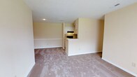 This single-floor residential unit offers a straightforward, functional layout designed to maximize usable space. The living area is open and airy, featuring wall-to-wall carpeting in a soft, neutral beige tone that adds warmth underfoot. The walls are painted in a light cream-yellow hue, contributing to a bright and welcoming ambiance throughout the space.

The kitchen is positioned at the rear of the open-concept living area and is defined by a peninsula counter that serves as a natural dividing element between the cooking and living zones. The kitchen is equipped with black appliances, including a range and an overhead microwave, and is complemented by white upper cabinetry that keeps the space feeling clean and organized. The countertops are light in color, maintaining the cohesive neutral palette of the unit.

The open layout between the kitchen and living space allows for flexible furniture arrangement and creates a sense of continuity throughout the floor. Recessed lighting in the kitchen area provides functional illumination, while the overall design prioritizes practicality. White baseboards run along the perimeter of the rooms, adding a subtle finishing detail. The space presents a clean, move-in ready environment suitable for a variety of living arrangements.
