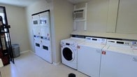 This shared laundry facility is designed for practical, everyday use and is outfitted with multiple commercial-grade machines to accommodate residents efficiently. The room features a set of stacked dryer units along one wall, maximizing vertical space, while front-loading washers and top-loading machines are positioned along the opposite side, providing users with a variety of laundry options. Each machine is numbered for easy identification. The space is climate-controlled with a wall-mounted air conditioning unit positioned above the machines, ensuring comfort during use. The room is finished with light-toned walls and tile flooring, contributing to a clean and well-maintained appearance. Adequate spacing between the machines allows for ease of movement and loading. The overall layout is straightforward and functional, prioritizing accessibility and efficiency. The facility is well-lit, creating a bright and hygienic environment suited for communal use.