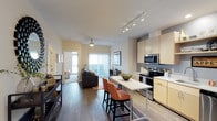 This single-floor residence offers a thoughtfully designed open-concept layout that seamlessly connects the kitchen, dining, and living areas into one cohesive and functional space. The kitchen is outfitted with light maple cabinetry, stainless steel appliances including a range and over-the-range microwave, and a movable island with a white countertop that doubles as a casual dining and prep surface. Two warm-toned bar stools provide additional seating at the island, adding a welcoming touch to the space. Hardwood-style flooring extends throughout the main living area, contributing to a clean and contemporary aesthetic. The living room benefits from abundant natural light streaming through large windows and sliding glass doors that open to an outdoor balcony, creating a bright and airy atmosphere. A ceiling fan overhead adds to the comfort of the space. Track lighting in the kitchen ensures excellent task illumination, while the open layout allows light to travel freely throughout. A hallway visible from the main area leads to additional rooms, including what appears to be a bedroom, further rounding out this well-appointed and efficiently arranged residence.