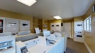 This shared laundry room is a well-organized, functional facility designed to serve multiple residents. The room features several top-loading washing machines, including Whirlpool units, arranged in a row along the floor, complemented by stacked dryer units mounted along the walls. The warm yellow-painted walls contribute a clean, utilitarian aesthetic, while overhead fluorescent lighting panels ensure the space is well-lit throughout. A tiled countertop area along one side of the room provides a convenient surface for sorting and folding laundry. The stacked dryers are built into wall alcoves, maximizing the efficient use of space. A bulletin board is mounted on the wall, serving as a communication point for residents. The room is finished with a durable hard-surface floor suitable for a high-traffic utility space. A door at the rear provides additional access to the facility. Overall, the laundry room is straightforward and practical, with all essential equipment clearly accessible and logically arranged to support ease of use for its residents.