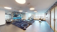 This well-appointed fitness studio at 3850 Boston St in Baltimore, Maryland offers a thoughtfully designed, single-floor layout built to support a wide range of workout styles. The main exercise area is generously sized, featuring durable interlocking rubber flooring and a large branded training mat at its center, ideal for functional and group fitness activities. Circular flush-mount lighting fixtures illuminate the space evenly, complementing the natural light that pours in through a bank of large windows along one wall, providing open views of the surrounding area. The studio is equipped with an impressive array of fitness equipment, including a treadmill, rowing machine, air bike, and weight training stations. A multi-tiered equipment rack stores medicine balls, resistance bands, TRX suspension trainers, foam rollers, and stability accessories, keeping the space organized and versatile. One wall features a decorative patterned wallcovering that adds a distinctive visual character, alongside a wall-mounted flat-screen television and a set of personal storage lockers for convenience. Frosted glass partition walls and doors define additional zones within the studio while maintaining an open, airy feel throughout. The clean white walls and modern finishes create a bright, motivating environment well-suited for both individual training and guided fitness sessions.