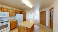 This single-floor residence offers a practical and comfortable layout designed for everyday living. The kitchen serves as a central hub of the home, featuring warm oak cabinetry, light countertops, a center island, and a full suite of white appliances including a refrigerator, electric range, microwave, and dishwasher. Tile flooring runs throughout the kitchen, which also benefits from natural light through a window with blinds and convenient access to an exterior door. The kitchen opens seamlessly into a generously sized living area, which is carpeted and fitted with a ceiling fan overhead and a pendant light fixture, creating a bright and airy atmosphere. Adjacent carpeted rooms serve as bedrooms, each featuring neutral walls, warm wood baseboards, and windows that bring in natural light and outdoor views. The bathroom is well-appointed with oak vanity cabinetry, a large mirror, a linen tower for additional storage, and tile flooring. A particularly convenient feature is the dedicated in-unit laundry area with a stacked washer and dryer unit, streamlining household routines. Throughout the space, consistent use of oak wood tones, neutral walls, and tile and carpet flooring creates a cohesive and functional living environment.