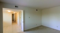 This single-floor residential space offers a straightforward and functional layout designed for comfortable everyday living. The main room features wall-to-wall carpet underfoot, contributing to a warm and quiet atmosphere, while the adjoining hallway transitions to easy-to-maintain tile flooring. The space is finished with smooth, neutral-toned walls and a textured ceiling, giving each room a clean and understated aesthetic.

Multiple interior doors visible from the main room provide access to additional spaces, which appear to include a closet and connecting rooms, suggesting a practical arrangement that supports organization and storage. Ceiling-mounted HVAC vents are present throughout, ensuring climate control across the space. Overhead lighting fixtures illuminate the hallway, adding to the overall brightness of the interior.

The open wall space in the main room offers flexibility in furniture arrangement, allowing the room to be configured to suit a variety of needs. The transition between the carpeted room and the tiled hallway creates a clear distinction between living and utility areas. Overall, this space presents a clean, move-in-ready environment with a neutral palette that serves as a versatile canvas for personalization.