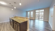 This thoughtfully designed 1-bedroom, 1-bathroom residence spans a single floor and offers a cohesive blend of modern finishes and functional living spaces. The open-concept kitchen and living area serves as the heart of the home, featuring rich dark-cabinet cabinetry, granite countertops, and a peninsula with pendant lighting overhead. Wood-look flooring runs throughout the main living area, complementing the sleek black stainless appliances, including a range, microwave, refrigerator, and dishwasher. Floor-to-ceiling sliding glass doors flood the living room with natural light and provide direct access to a private balcony, where scenic views of a nearby waterway and surrounding trees create a serene outdoor retreat. The spacious bedroom is finished with plush carpeting, a ceiling fan, and a window that continues the connection to the natural surroundings. A dedicated closet includes built-in wire shelving and conveniently houses a stacked washer and dryer. The full bathroom features a granite-topped dark vanity, a large mirror, and a tub-shower combination with tile surround. Neutral tones and clean architectural lines give the entire residence a bright, contemporary ambiance.