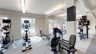 Located at 897–911 Axtell Dr in Cayce, South Carolina, this fitness center offers a thoughtfully designed, single-floor layout built to support a comprehensive workout experience. The facility is outfitted with a wide array of equipment, including cable machines, ellipticals, rowing machines, spin bikes, and a dedicated free weights area with kettlebell racks and a weight bench. Durable rubber-tile flooring covers the workout zones, providing stability and impact absorption throughout.

Large windows line the perimeter of the space, flooding the interior with abundant natural light and offering views of the surrounding outdoor area, including a visible pool and landscaping. The bright, airy atmosphere is complemented by modern linear lighting fixtures mounted to the ceiling, which features clean crown molding details. Full-length mirrored walls in multiple sections of the facility enhance the sense of openness and allow users to monitor their form during exercise.

The design incorporates accent walls in deep navy and charcoal tones, adding a polished, contemporary aesthetic. A dedicated hallway area features a dual-basin drinking fountain and bottle-filling station set against a textured stone tile wall. A restroom with wood-look flooring and accessible safety bars is also accessible from this corridor. The overall environment is clean, functional, and purpose-built for fitness.