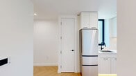 Located at 104 Fleet Pl in the heart of Downtown Brooklyn, this one-bedroom, one-bathroom residence offers a thoughtfully designed layout with a clean, contemporary aesthetic throughout. The open-plan living and kitchen area is anchored by striking herringbone-patterned hardwood floors that run seamlessly through the space, complemented by crisp white walls and recessed lighting that create a bright, airy ambiance.

The modern kitchen features sleek white cabinetry, open shelving, a stainless steel range, and a streamlined countertop with an undermount sink — an efficient and stylish space for cooking and entertaining. Black-framed hardware on the cabinetry and doors adds a bold, refined contrast to the otherwise light palette.

The bedroom is a serene retreat highlighted by dramatic floor-to-ceiling, black-framed windows that frame sweeping panoramic views of the Brooklyn skyline and beyond, flooding the room with natural light. The herringbone flooring continues here, maintaining a cohesive design language.

The bathroom is a standout feature, combining terrazzo flooring, vertical tile walls in warm neutral tones, a wood-framed vanity with black metal accents, and a globe pendant light for an elevated, design-forward finish. A hallway connects the rooms with ease, completing this well-appointed urban residence.