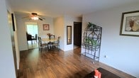 This single-floor residence offers a comfortable and functional living environment with a warm, inviting ambiance throughout. Rich hardwood flooring extends across the main living and dining areas, adding a sense of warmth and cohesion to the space. The open layout allows for easy flow between rooms, making the interior feel spacious and connected. The dining area is well-lit, benefiting from natural light streaming in through a nearby window, and is equipped with a ceiling fan that enhances both comfort and airflow. A round dining table with wrought iron chairs provides a classic, casual setting for meals. A full-length mirror is thoughtfully placed along one wall, adding depth to the space. Decorative touches, including a wrought iron baker's rack adorned with greenery, lend character and a homey feel to the interior. Neutral-toned walls throughout create a clean, versatile backdrop that complements the warm flooring. A hallway leads to additional rooms beyond, including what appears to be a closet or utility space with a dark-panel door. The overall design balances practicality with aesthetic appeal, making this a welcoming and livable single-floor home.