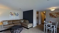 Located at 1600 Bonwood Road in Wilmington, Delaware, this single-floor residence offers a thoughtfully designed layout that balances comfort and functionality. The living area serves as the heart of the home, featuring plush wall-to-wall carpeting and a striking dark navy accent wall that adds a bold, modern contrast to the surrounding white walls. A large sectional sofa and a sleek circular coffee table anchor the space, creating an inviting area for relaxation. Colorful botanical artwork adorns the walls, adding a lively touch to the neutral palette. The open-concept design allows for a natural transition from the living room into the dining area, where a simple dining set is visible, ideal for everyday meals. Beyond the living space, a corridor leads to the kitchen, which is equipped with standard appliances and cabinetry, offering a practical and efficient workspace. The warm, ambient lighting throughout the hallways and adjoining rooms contributes to a cozy and welcoming atmosphere. The overall design of the space emphasizes clean lines, a cohesive color scheme, and a comfortable, livable environment well-suited for everyday living.