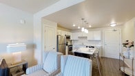 This single-floor residence offers a thoughtfully designed open-concept layout that seamlessly connects the living, dining, and kitchen areas into one cohesive and functional space. The kitchen is a standout feature, outfitted with crisp white cabinetry, a clean white countertop, and a full suite of stainless steel appliances including a refrigerator, microwave, and range. A set of elegant pendant lights hangs above the dining area, casting warm, focused light over the table and accentuating the kitchen's modern aesthetic. Recessed lighting throughout further brightens the space, complementing the soft, neutral tones of the walls and ceilings. The dining area comfortably accommodates a small table with contemporary clear-back chairs, blending style with practicality. Transitioning into the living area, light-colored striped upholstered chairs provide a cozy and inviting seating arrangement. Hardwood-style flooring runs throughout, adding warmth and continuity to the space. The overall ambiance is clean, airy, and welcoming, with a design sensibility that balances modern convenience with comfortable, everyday living.