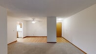 This single-floor residence offers a practical and comfortable layout with a clean, neutral aesthetic throughout. The open living area is defined by a transition from plush carpeting to light hardwood-style flooring, creating distinct zones within the space. A ceiling fan overhead adds both function and a classic touch, while a structural column adds architectural character and helps delineate the living and dining areas.

The kitchen features warm wood cabinetry with ample counter space in a light laminate finish, and includes a white refrigerator and range. The hardwood-style flooring continues here, contributing to a cohesive feel.

The bedrooms are generously proportioned, each finished with soft wall-to-wall carpeting and large windows dressed with vertical blinds, allowing natural light to filter in while offering views of the surrounding greenery. Baseboard heating runs along the perimeter, and built-in closets with warm wood-toned doors provide practical storage. One bedroom features a wall-mounted air conditioning unit for added comfort.

The bathroom is well-appointed with a corner shower stall with frosted glass enclosure, a vanity with wood cabinetry and a wide mirror, and classic tile flooring. Textured ceilings are consistent throughout, completing the straightforward and functional character of this residence.