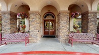 This single-story space makes a striking first impression with its covered entryway, where stacked stone columns flank a set of arched double doors with decorative ironwork. Hanging flower baskets add vibrant color to the exterior, and a pair of benches provide a welcoming spot to pause before entering.

Inside, the focal point of the main lounge is a commanding floor-to-ceiling stacked stone fireplace, adorned with candelabras and decorative metalwork above the mantel. The warm, earthy tones of the stonework are complemented by exposed wood ceiling beams that lend a rustic yet refined character to the space. Ornate wrought-iron wall accents and lush greenery placed at height add visual depth and warmth throughout.

The seating area is generously arranged with floral-patterned sofas and armchairs accented by jewel-toned throw pillows, centered around a glass-top coffee table. Arched doorways on either side of the fireplace lead to additional areas, hinting at a broader layout that includes dining and lounge spaces. The overall ambiance is rich, cozy, and welcoming, with a Mediterranean-inspired aesthetic throughout.