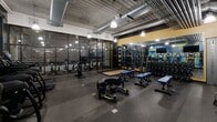 Located at 619 E University Ave in Ann Arbor, Michigan, this fitness facility offers a comprehensive workout environment across a single, open floor. The space is designed with an industrial aesthetic, featuring exposed ductwork and ventilation on the ceiling, pendant lighting, and durable rubber flooring throughout — all contributing to a functional and energetic atmosphere.

The strength training area is well-appointed with a Matrix power rack, a full dumbbell rack, adjustable blue benches, weight plates, and wall-to-wall mirrors that enhance the sense of openness and allow users to monitor their form. A dedicated functional training zone includes a heavy tire, a punching bag, and resistance-based equipment, catering to a variety of training styles.

The cardio section features a row of upright bikes and stair-climber machines, positioned near a bold, colorful branded mural in blue and yellow that adds a dynamic visual focal point to the space. A cable machine station and additional free weights round out the training zones.

Near the entrance, a snack and beverage vending machine provides convenient refreshment access. Large glass partitions along one wall offer a view into an adjacent landscaped area, adding a subtle natural element to the otherwise industrial setting. The layout is efficient, organized, and designed to support a wide range of fitness activities.
