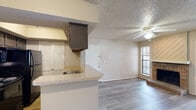 This single-floor residence offers a well-designed, open-concept layout that seamlessly connects the kitchen and living areas. The kitchen is equipped with dark cabinetry, black appliances including a range, microwave, refrigerator, and dishwasher, and features a light-toned countertop peninsula with a built-in sink that opens to the main living space. Louvered bi-fold doors provide convenient storage access nearby. The living area is bright and welcoming, highlighted by a charming brick fireplace with a shiplap wood surround that serves as a focal point. A ceiling fan overhead and a set of French doors allow natural light to fill the space and provide outdoor access. Durable wood-look flooring flows consistently throughout, complemented by textured ceilings that add a classic touch. The bedroom is a comfortable, well-proportioned room with the same flooring and neutral wall tones, offering a clean and versatile canvas. The bathroom is thoughtfully appointed with a wide vanity featuring dark cabinetry, a light countertop, a single sink, a large mirror with overhead lighting, and a tub-shower combination. The overall ambiance of the space is clean, functional, and move-in ready.