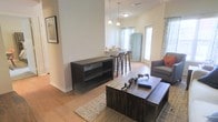 This single-floor space showcases an open-concept design that seamlessly integrates the living and dining areas into one cohesive, functional layout. The living area features warm hardwood-style flooring that extends throughout, anchored by a woven area rug and complemented by a rustic dark-wood coffee table and matching media console. A comfortable sofa with accent pillows adds a welcoming touch to the space.

The layout flows naturally toward a casual dining area, where a white bar-height counter is paired with wooden barstools, creating an ideal spot for casual meals or conversation. Pendant lights hang above, adding both task lighting and a stylish focal point. Natural light pours in through multiple windows dressed with patterned curtains, contributing to the airy, open ambiance of the space.

A distinctive teal-painted storage cabinet near the entry adds a charming, vintage-inspired accent. The walls are kept in a clean, neutral tone, allowing the warm wood tones and carefully chosen furnishings to stand out. Overall, the space strikes a balance between casual comfort and thoughtful design, making it both practical and inviting.