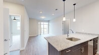 This well-appointed single-floor residence showcases a thoughtfully designed open-concept layout that seamlessly connects the kitchen and living areas. The kitchen serves as a central highlight, featuring a large granite-topped island with an undermount sink, sleek dark cabinetry, stainless steel appliances including a refrigerator, range, and microwave, and stylish pendant lighting overhead. Light-toned wood-style flooring flows throughout the main living areas, complementing the neutral color palette of the walls and creating a bright, airy atmosphere.

The living area extends toward a private balcony accessible through full glass sliding doors, inviting abundant natural light and outdoor views into the space. Bedrooms offer plush carpeting underfoot, generous proportions, and access to a walk-in closet outfitted with built-in shelving and hanging systems. One bedroom includes a window with elevated views of the surrounding area, adding a sense of openness. The bathroom is finished with matching granite countertops, an undermount sink, dark-framed mirror, and modern fixtures, maintaining the consistent upscale aesthetic found throughout the residence. Recessed lighting and clean white trim complete the polished, contemporary interior design.