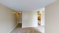 This single-floor unit features two bedrooms and one bathroom within a well-utilized floor plan. The living area is finished with light-toned carpeting and neutral cream-colored walls, creating a bright, clean atmosphere. A textured ceiling adds subtle character to the space, while tile flooring near the entryway provides a durable transition zone at the front door. Ceiling-mounted HVAC vents are integrated throughout, ensuring consistent climate control. The kitchen is positioned adjacent to the main living area, partially separated by a dividing wall that defines each zone without fully closing them off. The kitchen is outfitted with white cabinetry, a countertop workspace, and tile flooring, offering a functional cooking environment. The entryway, visible from the living area, provides a straightforward entry point into the unit. The overall design prioritizes practicality and ease of use, with a neutral color palette throughout that allows for flexible interior styling. The two bedrooms offer dedicated sleeping spaces, complementing the shared living and kitchen areas for a complete residential layout.