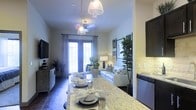 This thoughtfully designed single-floor space offers a seamless open-plan layout that connects the kitchen, dining, and living areas into one cohesive and inviting environment. The kitchen island takes center stage, featuring striking granite countertops and serving as both a functional workspace and a dining area, complete with elegant place settings that highlight its versatility.

Overhead, a pair of decorative pendant lights with ornate shades cast a warm, ambient glow across the island, complementing the overall lighting scheme throughout the space. Dark hardwood flooring grounds the interior and adds a rich contrast to the light-toned walls and furnishings.

The living area is anchored by a clean-lined sofa accented with patterned throw pillows, accompanied by lush indoor plants — including a prominent fiddle-leaf fig and a sculptural cactus arrangement — that bring life and texture to the space. A flat-screen television is mounted on the far wall, adding a modern touch.

At the far end of the room, French doors framed by sheer curtains allow abundant natural light to pour in, creating a bright and airy atmosphere. The ceiling height adds to the sense of openness, making this a well-appointed and welcoming space ideal for both everyday living and entertaining.