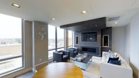 This single-floor space showcases a sophisticated, contemporary design centered around an open-concept living area. The focal point of the room is a dark wood-paneled entertainment wall, which houses a wall-mounted flat-screen television above a sleek linear fireplace, creating a dramatic and functional feature. Recessed lighting in the white ceiling complements the natural light that floods the space through expansive floor-to-ceiling windows, which frame impressive elevated city views.

The living area is thoughtfully furnished with a pair of charcoal upholstered armchairs and a white sofa, arranged around a low-profile rectangular coffee table atop a neutral area rug, creating an inviting yet refined seating arrangement. A round warm-toned wood table is partially visible, suggesting a dedicated dining area nearby.

The color palette throughout is understated and cohesive, combining warm taupe walls, crisp white ceilings, and dark wood accents. A decorative white stag head wall mount adds a subtle artistic touch. Built-in shelving on the entertainment wall provides both display and storage opportunities, contributing to the space's clean, organized aesthetic.