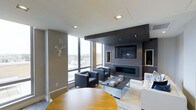 This single-floor space showcases a sophisticated, contemporary design centered around an open-concept living area. The focal point of the room is a dark wood-paneled entertainment wall, which houses a wall-mounted flat-screen television above a sleek linear fireplace, creating a dramatic and functional feature. Recessed lighting in the white ceiling complements the natural light that floods the space through expansive floor-to-ceiling windows, which frame impressive elevated city views.

The living area is thoughtfully furnished with a pair of charcoal upholstered armchairs and a white sofa, arranged around a low-profile rectangular coffee table atop a neutral area rug, creating an inviting yet refined seating arrangement. A round warm-toned wood table is partially visible, suggesting a dedicated dining area nearby.

The color palette throughout is understated and cohesive, combining warm taupe walls, crisp white ceilings, and dark wood accents. A decorative white stag head wall mount adds a subtle artistic touch. Built-in shelving on the entertainment wall provides both display and storage opportunities, contributing to the space's clean, organized aesthetic.
