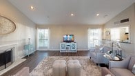 This single-floor home offers a thoughtfully designed open-concept layout that seamlessly connects the kitchen and living areas. The living room is a standout space, showcasing soaring vaulted ceilings with recessed lighting that enhance the sense of openness and airiness throughout. Rich, dark hardwood flooring runs across the main living area, providing a warm contrast to the neutral, light-toned walls.

A classic fireplace with a white brick surround serves as a focal point in the living room, complemented by decorative wall art above it. Large windows fitted with blinds and white curtains allow an abundance of natural light to fill the space while offering views of the surrounding exterior. The living area is generously proportioned, easily accommodating multiple seating arrangements.

The kitchen connects to the living space via a pass-through counter, featuring bold dark granite countertops with a double stainless steel sink. White cabinetry and bar-height seating add a clean, functional touch. The overall aesthetic throughout the home is cohesive and contemporary, blending neutral tones with tasteful accents, creating an inviting and well-appointed atmosphere ideal for both everyday living and entertaining.