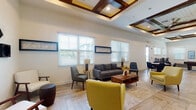 This single-story community space is designed to offer a welcoming and versatile environment for socializing and relaxation. The interior is unified by light wood-look herringbone-patterned flooring that runs throughout, complementing the crisp white walls and creating a clean, contemporary aesthetic.

The main lounge area serves as the centerpiece of the space, featuring an elegant coffered ceiling with dark wood accents and recessed lighting that adds architectural depth. A curated mix of mid-century modern furniture, including a gray sofa, yellow accent chairs, and white armchairs, is arranged around a rustic wood coffee table. Multiple windows with white horizontal blinds flood the area with natural light, while framed blue wall art adds tasteful decorative touches.

Adjacent to the lounge, a kitchenette is outfitted with white cabinetry, granite countertops, a tile backsplash, stainless steel appliances, and open floating shelves. A curved island with bar stools makes it a practical gathering point.

A casual dining nook features high-top tables and industrial-style bar stools, providing a relaxed setting for informal meetings or socializing. Direct exterior access is available via a glass-panel door, connecting the interior to the outdoors seamlessly.