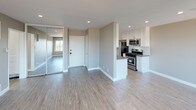 This single-floor unit offers a clean, modern aesthetic throughout, characterized by neutral taupe and gray tones, recessed lighting, and a cohesive mix of hard and soft flooring. The open-concept living and dining area is bright and airy, featuring light wood-look vinyl flooring, a wall-mounted air conditioning unit, and large sliding glass doors that open onto a private balcony with outdoor views. An oversized window further fills the space with natural light, creating an inviting atmosphere. The kitchen is efficiently laid out with white shaker-style cabinetry, dark countertops, a stainless steel sink, and a full suite of stainless steel appliances including a gas range, microwave, and refrigerator. Adjacent to the main living area, the bedroom offers plush carpeting underfoot and a floor-to-ceiling mirrored sliding closet that enhances the sense of space. A hallway connects the bedroom to a bathroom featuring a combined tub and shower with brushed nickel fixtures, wood-look tile flooring, and a vanity with dark countertop and white cabinetry. Throughout the unit, clean white baseboards and smooth walls contribute to a polished, move-in-ready finish.