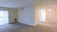 This single-floor residence offers a straightforward, functional layout designed for comfortable everyday living. The main living area is finished with wall-to-wall carpeting in a neutral beige tone, creating a warm and cohesive feel throughout the space. The room is defined by clean white walls and a flat ceiling with integrated HVAC vents, ensuring climate control throughout. A built-in shelving unit is recessed into one wall of the living area, providing convenient storage or display space without encroaching on the room's open footprint. A wide hallway extends from the main living space, bathed in natural light that brightens the interior and draws the eye toward the private areas of the home. Multiple doorways line the corridor, suggesting a series of rooms — likely bedrooms or utility spaces — arranged for accessibility and privacy. The transition between the carpeted living area and the hallway is clearly defined, maintaining a sense of organized flow between the common and private zones. Overall, the space presents a clean, move-in-ready canvas with a neutral palette and practical layout well-suited for a variety of living arrangements.