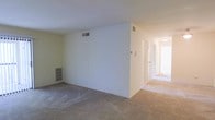 This single-floor residence offers a straightforward, functional layout designed for comfortable everyday living. The main living area is finished with wall-to-wall carpeting in a neutral beige tone, creating a warm and cohesive feel throughout the space. The room is defined by clean white walls and a flat ceiling with integrated HVAC vents, ensuring climate control throughout. A built-in shelving unit is recessed into one wall of the living area, providing convenient storage or display space without encroaching on the room's open footprint. A wide hallway extends from the main living space, bathed in natural light that brightens the interior and draws the eye toward the private areas of the home. Multiple doorways line the corridor, suggesting a series of rooms — likely bedrooms or utility spaces — arranged for accessibility and privacy. The transition between the carpeted living area and the hallway is clearly defined, maintaining a sense of organized flow between the common and private zones. Overall, the space presents a clean, move-in-ready canvas with a neutral palette and practical layout well-suited for a variety of living arrangements.