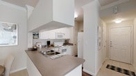 This single-floor residence features two bedrooms and two bathrooms, offering a thoughtfully designed layout ideal for comfortable everyday living. The kitchen serves as a central highlight, showcasing white cabinetry that extends to the upper walls, providing ample storage space. A large overhead range hood anchors the cooking area, which includes a white electric range, built-in microwave, and a full-size refrigerator. The countertops, finished in a neutral gray tone, extend into a peninsula that doubles as a breakfast bar, creating a natural division between the kitchen and the adjacent living area. A double-basin sink with a modern faucet is integrated into the peninsula for added convenience. Open shelving near the kitchen displays decorative items, adding personality to the space. Crown molding is visible along the ceiling, lending an elevated, refined touch to the interior. The hallway visible beyond the kitchen leads to the bedrooms and bathrooms, with neutral-toned carpeting and light walls creating a clean, cohesive aesthetic throughout. The overall ambiance is bright and welcoming, with white finishes and neutral tones contributing to an open, airy feel.