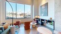 Located at 3750 Blake St in Denver's dynamic RiNo neighborhood, this single-floor lobby space combines contemporary design with warm, inviting elements throughout. Polished concrete floors extend across the entire floor plan, reflecting light and lending an industrial-modern aesthetic that sets the tone upon entry. Large black-framed glass doors and expansive windows flood the interior with abundant natural light while offering views of the surrounding cityscape.

The main entrance corridor features striking geometric pendant lighting and open sightlines that draw visitors deeper into the space. Exposed concrete columns serve as bold architectural anchors, contrasting with crisp white walls and warm wood base trim. A built-in gas fireplace with a sleek white surround anchors a cozy lounge area appointed with teal and rust-toned seating, a glass coffee table, and a potted fiddle-leaf fig tree.

A reading nook nearby offers additional seating centered around a curated built-in shelving unit displaying books and décor, accented by large-scale portrait artwork that adds cultural character. An open event or multipurpose area features high ceilings with recessed lighting, a projection screen, and gallery-style art installations on the walls. Throughout the space, thoughtfully selected furnishings and greenery create a welcoming, community-oriented atmosphere well-suited for gathering, working, or relaxing.