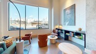 Located at 3750 Blake St in Denver's dynamic RiNo neighborhood, this single-floor lobby space combines contemporary design with warm, inviting elements throughout. Polished concrete floors extend across the entire floor plan, reflecting light and lending an industrial-modern aesthetic that sets the tone upon entry. Large black-framed glass doors and expansive windows flood the interior with abundant natural light while offering views of the surrounding cityscape.

The main entrance corridor features striking geometric pendant lighting and open sightlines that draw visitors deeper into the space. Exposed concrete columns serve as bold architectural anchors, contrasting with crisp white walls and warm wood base trim. A built-in gas fireplace with a sleek white surround anchors a cozy lounge area appointed with teal and rust-toned seating, a glass coffee table, and a potted fiddle-leaf fig tree.

A reading nook nearby offers additional seating centered around a curated built-in shelving unit displaying books and décor, accented by large-scale portrait artwork that adds cultural character. An open event or multipurpose area features high ceilings with recessed lighting, a projection screen, and gallery-style art installations on the walls. Throughout the space, thoughtfully selected furnishings and greenery create a welcoming, community-oriented atmosphere well-suited for gathering, working, or relaxing.