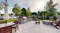 This expansive outdoor amenity space is thoughtfully designed to offer a refined yet relaxed setting for outdoor living and entertaining. The patio is finished with elegant paver stonework throughout, creating a cohesive and polished aesthetic underfoot. At the heart of the space sits a stone-clad circular fire pit surrounded by rich mahogany Adirondack chairs, providing a natural gathering point for relaxation. A built-in outdoor kitchen features a full-size stainless steel grill set into a handsome natural stone surround with ample counter space, making it ideal for al fresco dining. Multiple seating arrangements, including umbrella-shaded dining tables and additional Adirondack chairs, are distributed across the patio to accommodate a variety of group sizes. Low stone retaining walls with column accents define the perimeter, while carefully maintained landscaping—including young trees, evergreen shrubs, and manicured lawns—adds greenery and a sense of privacy. Classic black lamp posts punctuate the space, offering ambient lighting. The building's exterior, finished in gray siding and stacked stone veneer, provides an attractive architectural backdrop. A welcoming entry pathway with well-tended gardens leads to the building entrance, completing this well-appointed outdoor retreat.