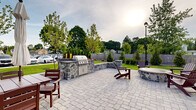 This expansive outdoor amenity space is thoughtfully designed to offer a refined yet relaxed setting for outdoor living and entertaining. The patio is finished with elegant paver stonework throughout, creating a cohesive and polished aesthetic underfoot. At the heart of the space sits a stone-clad circular fire pit surrounded by rich mahogany Adirondack chairs, providing a natural gathering point for relaxation. A built-in outdoor kitchen features a full-size stainless steel grill set into a handsome natural stone surround with ample counter space, making it ideal for al fresco dining. Multiple seating arrangements, including umbrella-shaded dining tables and additional Adirondack chairs, are distributed across the patio to accommodate a variety of group sizes. Low stone retaining walls with column accents define the perimeter, while carefully maintained landscaping—including young trees, evergreen shrubs, and manicured lawns—adds greenery and a sense of privacy. Classic black lamp posts punctuate the space, offering ambient lighting. The building's exterior, finished in gray siding and stacked stone veneer, provides an attractive architectural backdrop. A welcoming entry pathway with well-tended gardens leads to the building entrance, completing this well-appointed outdoor retreat.