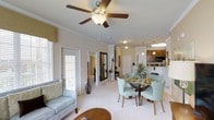 Located at 14411 Calloway Gap Rd in Raleigh, NC, this single-floor residence offers a thoughtfully designed open-concept layout that blends comfort and functionality. The living area is bright and inviting, featuring plush carpeting, a ceiling fan with light fixture, and large windows that welcome abundant natural light, creating a warm and airy atmosphere throughout.

The dining area flows naturally from the living space, anchored by a round glass-top table surrounded by light teal upholstered chairs, adding a touch of contemporary elegance. Decorative pendant lighting overhead enhances the dining experience and contributes to the home's refined aesthetic.

The kitchen is seamlessly integrated into the open floor plan, featuring dark wood cabinetry, light countertops, and a breakfast bar with pendant lighting, making it both practical and visually appealing. Built-in appliances, including a microwave and oven, add to the kitchen's functionality.

Architectural details such as columned transitions between spaces, vaulted ceilings, and a neutral color palette give the home a polished yet welcoming character. Additional rooms visible through the hallway suggest well-proportioned private spaces, completing this cohesive and comfortable single-level home.