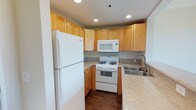 This single-floor space includes a well-appointed kitchen designed for practical everyday use. The kitchen features warm-toned, honey maple cabinetry that extends to the upper walls, providing generous storage throughout. Granite-style countertops in a neutral speckled finish offer durable workspace and complement the cabinetry's warm tones. White appliances — including a full-size refrigerator, gas range with oven, over-the-range microwave, and a double-basin sink with a modern arched faucet — are neatly integrated into an efficient L-shaped layout. Recessed lighting in the ceiling ensures the workspace is well-lit. The dark hardwood-style flooring anchors the kitchen and adds contrast to the lighter cabinetry and countertops. The overall design is clean and cohesive, with a functional aesthetic that balances practicality and style. The neutral color palette throughout creates a bright, welcoming atmosphere well-suited for cooking and everyday living.