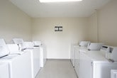 This shared laundry facility is a well-organized, single-floor space designed to accommodate the laundry needs of multiple users efficiently. The room features multiple commercial-grade Maytag top-load washing machines and dryers arranged along opposing walls, maximizing the use of available space while allowing easy access to each unit. The washers are coin-operated, providing a straightforward and convenient payment method for users.

The interior is finished with neutral, light beige walls and light-toned tile flooring, creating a clean and functional environment. The layout places washers on one side and dryers on the other, streamlining the laundry process. A centrally located door with a multi-pane window allows natural light to enter the room and provides a view of a green outdoor area, adding a pleasant touch to an otherwise utilitarian space.

Overhead ventilation ensures proper air circulation throughout the facility. Posted instructional and usage guideline notices are visible on the machines and walls, keeping users informed of proper operation. The space is straightforward in design, prioritizing practicality and ease of use, making it a reliable shared amenity for residents or occupants of the surrounding property.