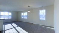 This single-floor space presents a clean, move-in-ready layout designed with simplicity and functionality in mind. The main living area is generously proportioned, offering an open and airy feel enhanced by soft, neutral wall tones that complement the plush wall-to-wall carpeting underfoot. Natural light floods the interior through multiple large, white-framed windows positioned along the exterior walls, casting bright patterns across the floor and creating a warm, inviting atmosphere throughout the day. A classic pendant light fixture hangs centrally from the smooth ceiling, providing a focal point and supplemental illumination that adds a touch of understated elegance to the space. The windows also provide views of the surrounding exterior environment, connecting the interior to the outside world. White baseboards and clean wall finishes contribute to the overall fresh and well-maintained character of the space. The open plan layout offers flexibility for a variety of furniture arrangements and uses, making it a versatile and practical environment. The combination of ample natural light, neutral finishes, and quality flooring gives this space a comfortable and livable quality that is both functional and visually appealing.